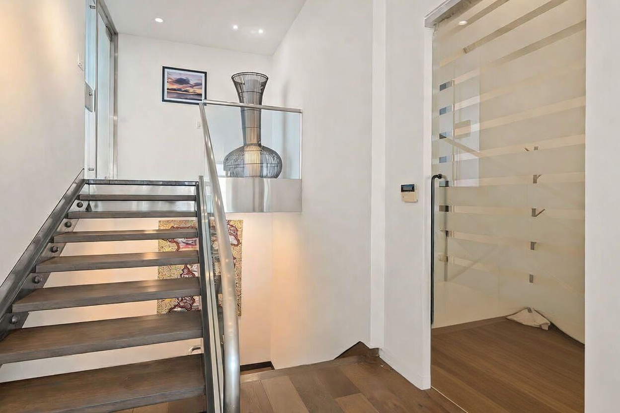 This modern entryway features a striking staircase with dark wood treads and a sleek metal railing. To the right, a frosted glass door with horizontal detailing provides privacy while allowing light to filter through, and a decorative vase sits atop a glass-railed landing. The space is characterized by clean lines, neutral walls, and a contemporary aesthetic that creates a sophisticated first impression.
