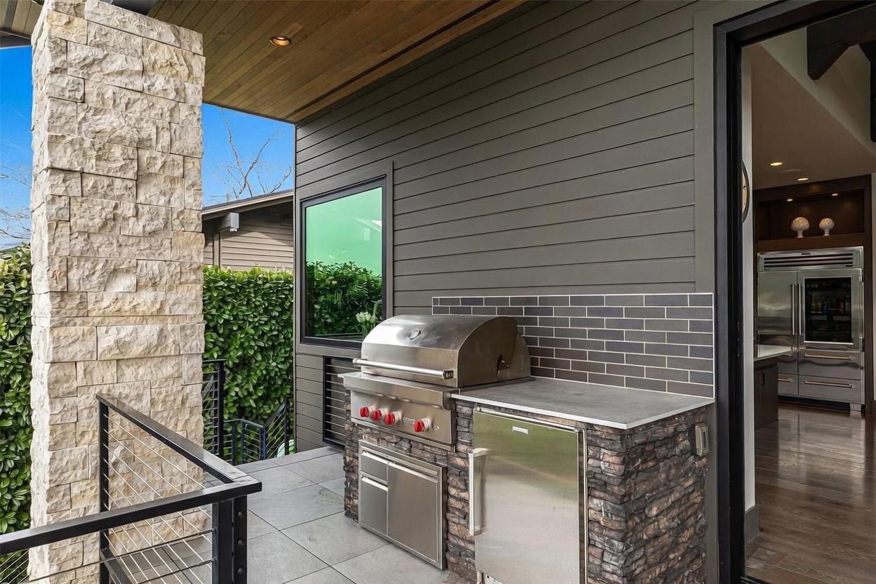This outdoor living space features a built-in stainless steel grill station with a stone base and a gray tiled backsplash, set against a dark horizontal-sided wall. A metal railing overlooks a landscaped area, while a large window provides a view into the interior kitchen space. The area is covered by a wood-paneled ceiling, creating a sophisticated and functional transition between indoor and outdoor entertaining zones.