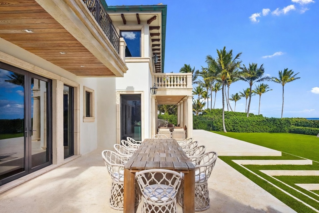 This elegant outdoor patio features a long wooden dining table surrounded by woven chairs, set against a bright, Mediterranean-inspired home exterior. The space includes a covered area with stone columns and a balcony above, transitioning seamlessly into a manicured lawn and lush tropical landscaping. The perspective captures the inviting al fresco dining area with a clear view of the expansive backyard and blue sky.