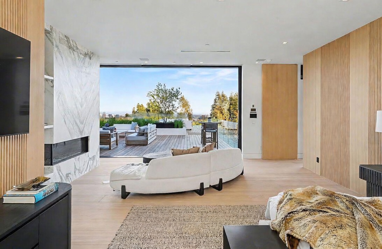This contemporary living space features light wood flooring and a prominent stone-clad chimney breast housing a linear fireplace. A curved white sofa serves as the centerpiece, positioned to face both the interior fireplace and the expansive outdoor deck visible through large sliding glass doors. The room transitions seamlessly to the exterior, creating an open-concept indoor-outdoor living experience with a view of the surrounding landscape.