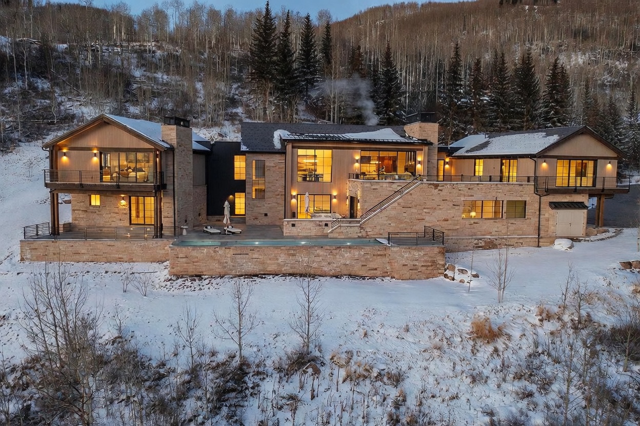This expansive mountain estate features a sophisticated blend of natural stone and wood siding, set against a backdrop of snow-covered trees. The multi-level design includes a prominent swimming pool on the terrace, multiple balconies, and large windows that illuminate the interior. The aerial perspective captures the home's integration into the rugged, wintry landscape, emphasizing its luxury and secluded setting.