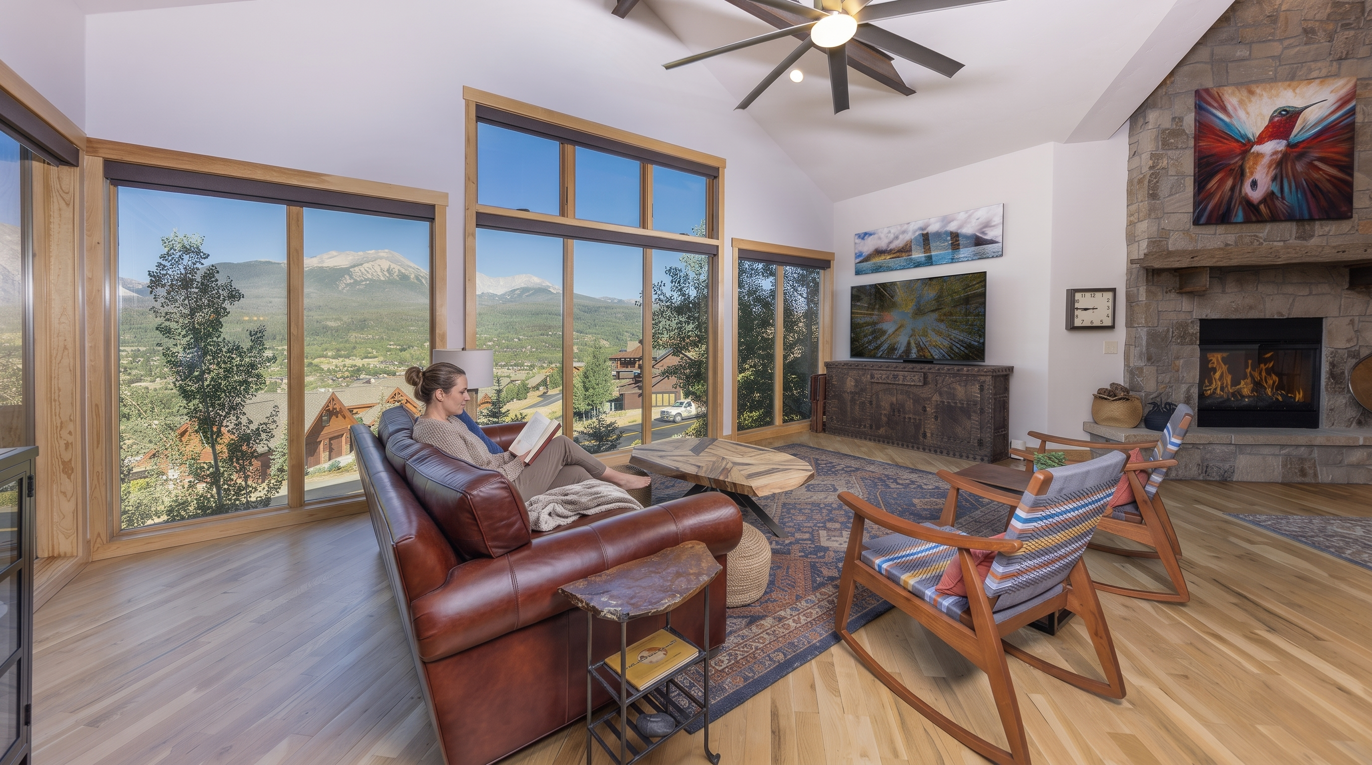 This spacious and sunlit living room features vaulted ceilings, rich hardwood flooring, and a stunning stone fireplace that serves as a focal point. Large floor-to-ceiling windows provide breathtaking panoramic views of the surrounding mountains and pine trees, creating an inviting atmosphere that blends indoor comfort with nature. The room is elegantly furnished with a leather sofa, two wooden rocking chairs, and an artisanal coffee table, all set upon a decorative area rug.
