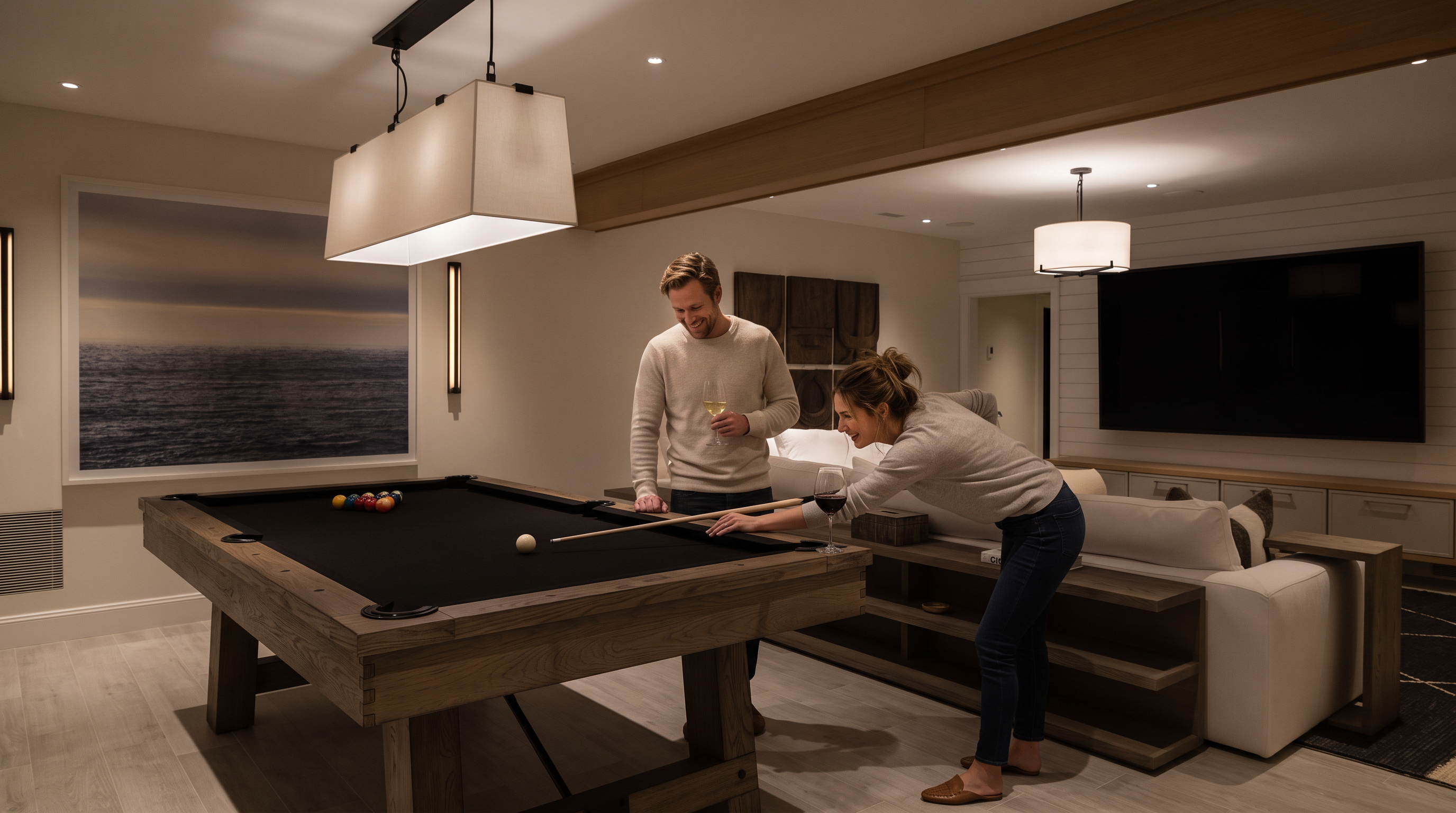 This spacious, modern basement recreation area features a pool table in the foreground and a comfortable lounge space with a large white sectional sofa facing a wall-mounted television. The room is accented by light wood tones, neutral walls, and decorative wall art, creating a cohesive and inviting atmosphere. The perspective captures the open-concept layout, highlighting the seamless transition between the gaming and media zones.