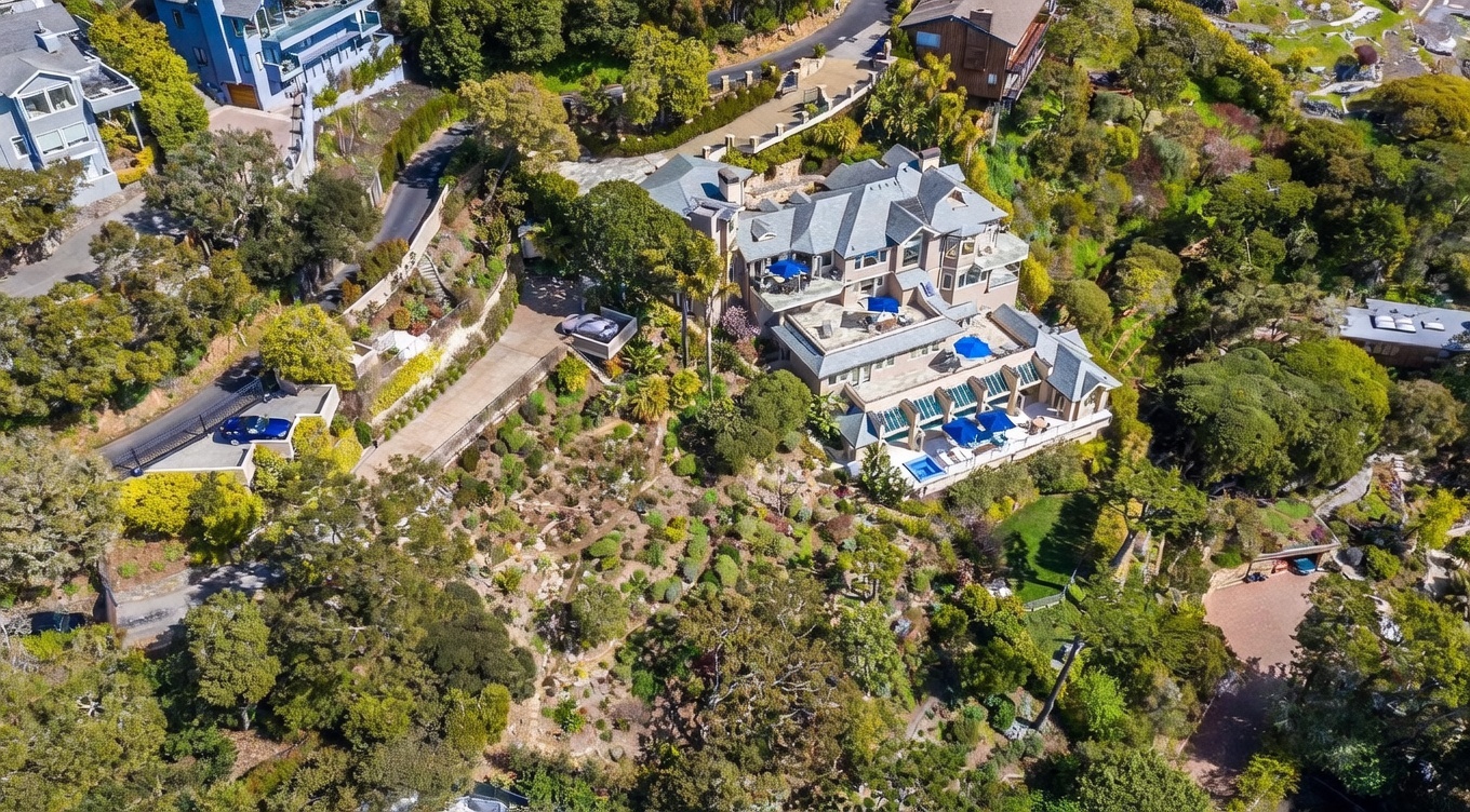This high-angle aerial view captures a sprawling, multi-level estate nestled into a lush, sloped hillside. The property features extensive terraced outdoor living spaces, including multiple patios with blue umbrellas, a swimming pool, and a winding driveway that leads to a detached garage. The surrounding landscape is densely wooded, emphasizing the home's secluded and private setting.