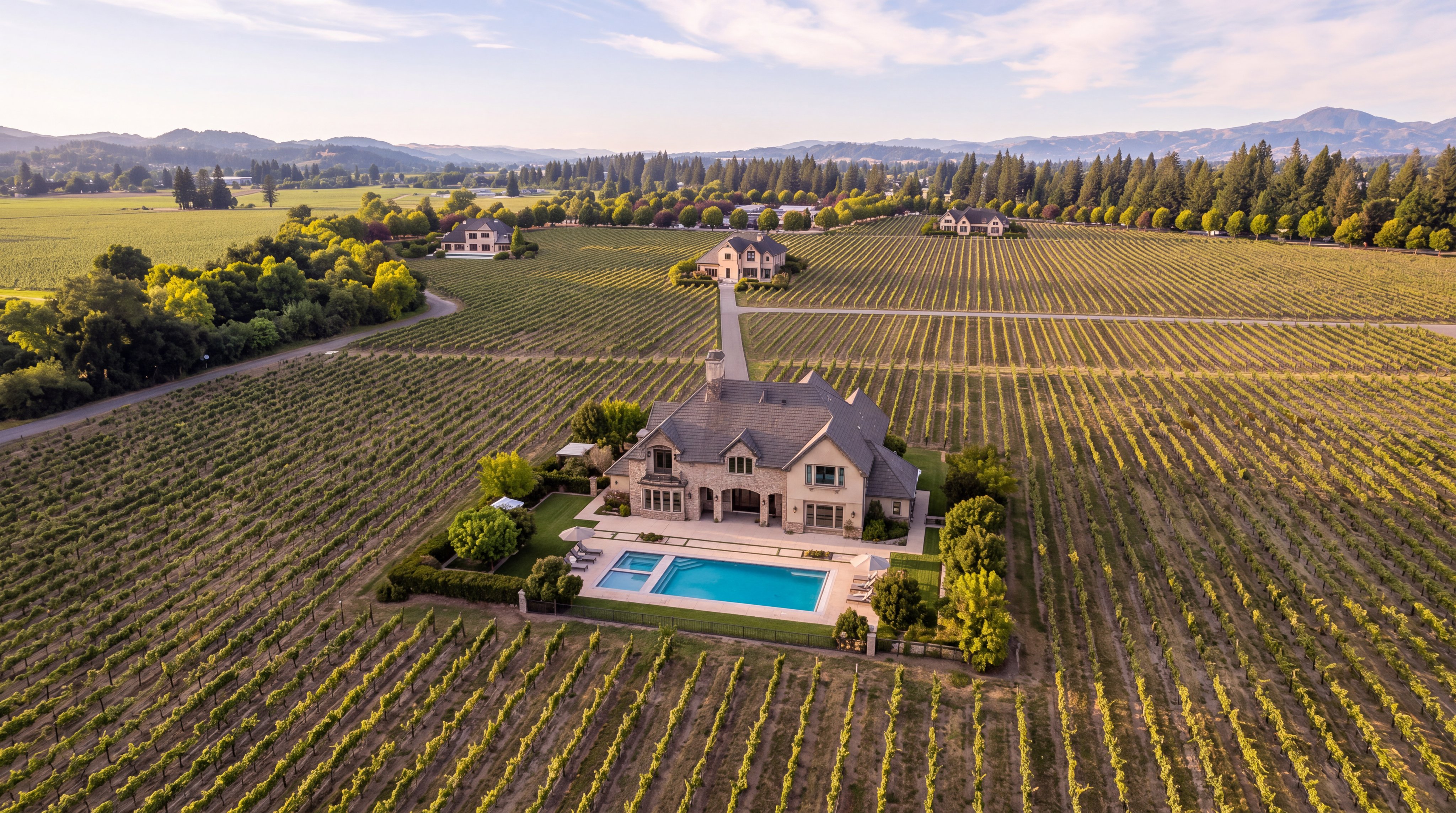 This stunning aerial view captures a luxurious estate surrounded by expansive, manicured vineyards. The property features a large multi-story residence with a classic architectural design, complemented by an inviting swimming pool and well-landscaped yard. The serene rural setting, framed by rolling hills in the distance, creates an atmosphere of elegance, privacy, and upscale countryside living.