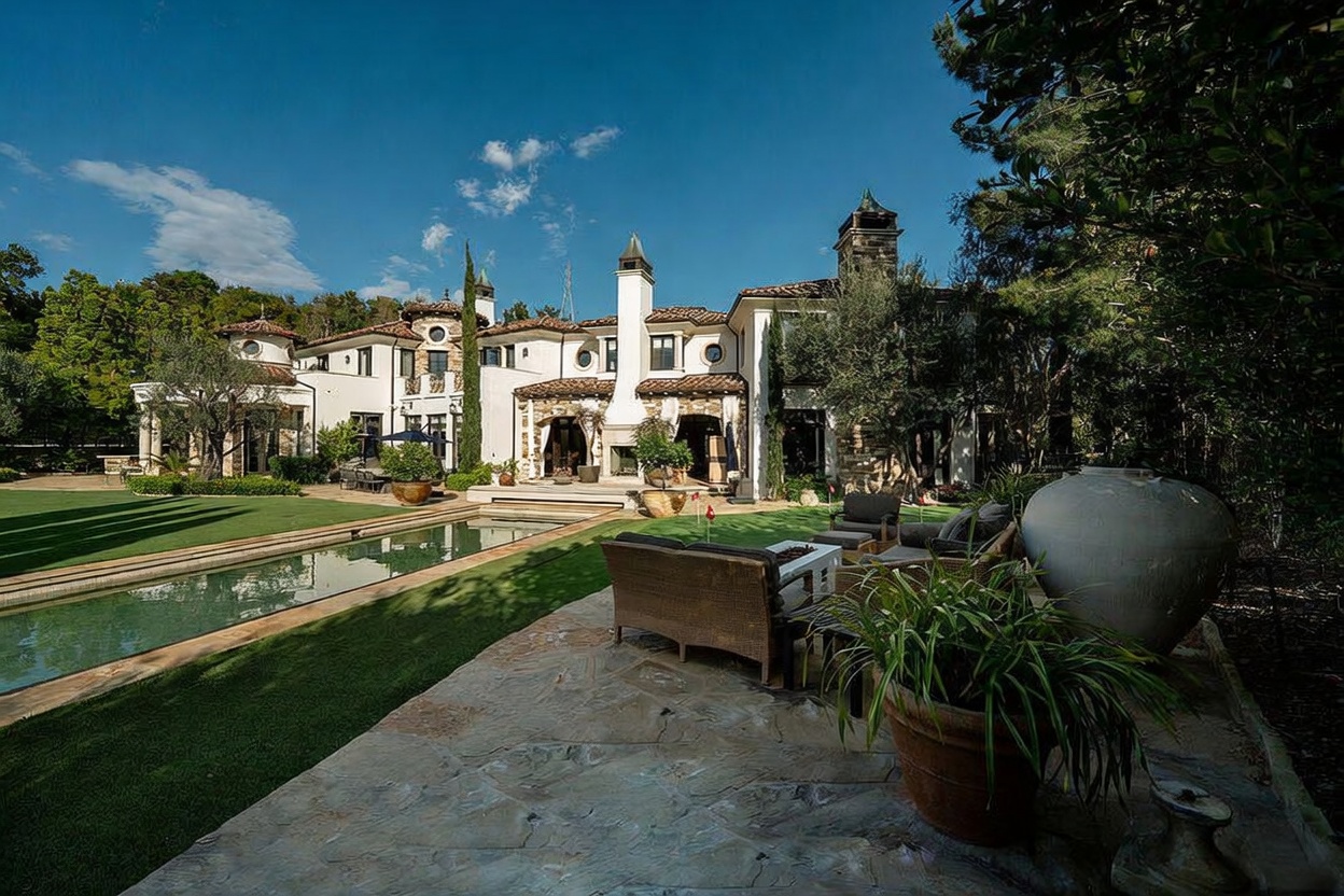 This expansive Mediterranean-style estate features a sprawling backyard with a long, rectangular swimming pool and a manicured lawn. The rear facade showcases intricate architectural details, including a prominent stone-clad chimney breast, arched doorways, and a stone patio furnished with comfortable outdoor seating. The scene is captured from a slightly elevated perspective, emphasizing the seamless transition between the luxurious indoor living spaces and the serene, landscaped outdoor environment.