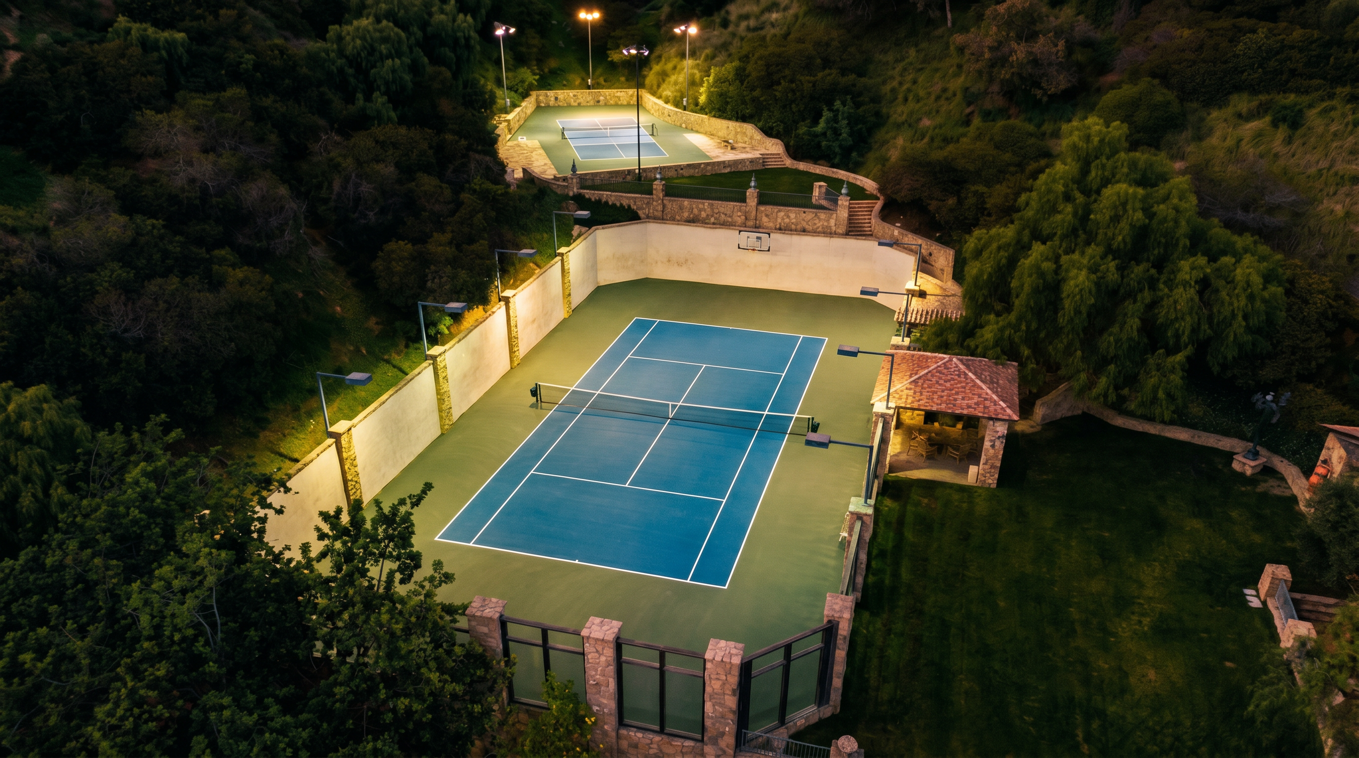 This high-angle aerial shot captures a luxurious private estate featuring two tiered tennis courts nestled into a lush, hillside landscape. The primary court is a vibrant blue and green hard court, complemented by a charming stone-roofed gazebo and surrounding manicured greenery. The perspective emphasizes the property's secluded, resort-like atmosphere and its integration into the natural terrain.