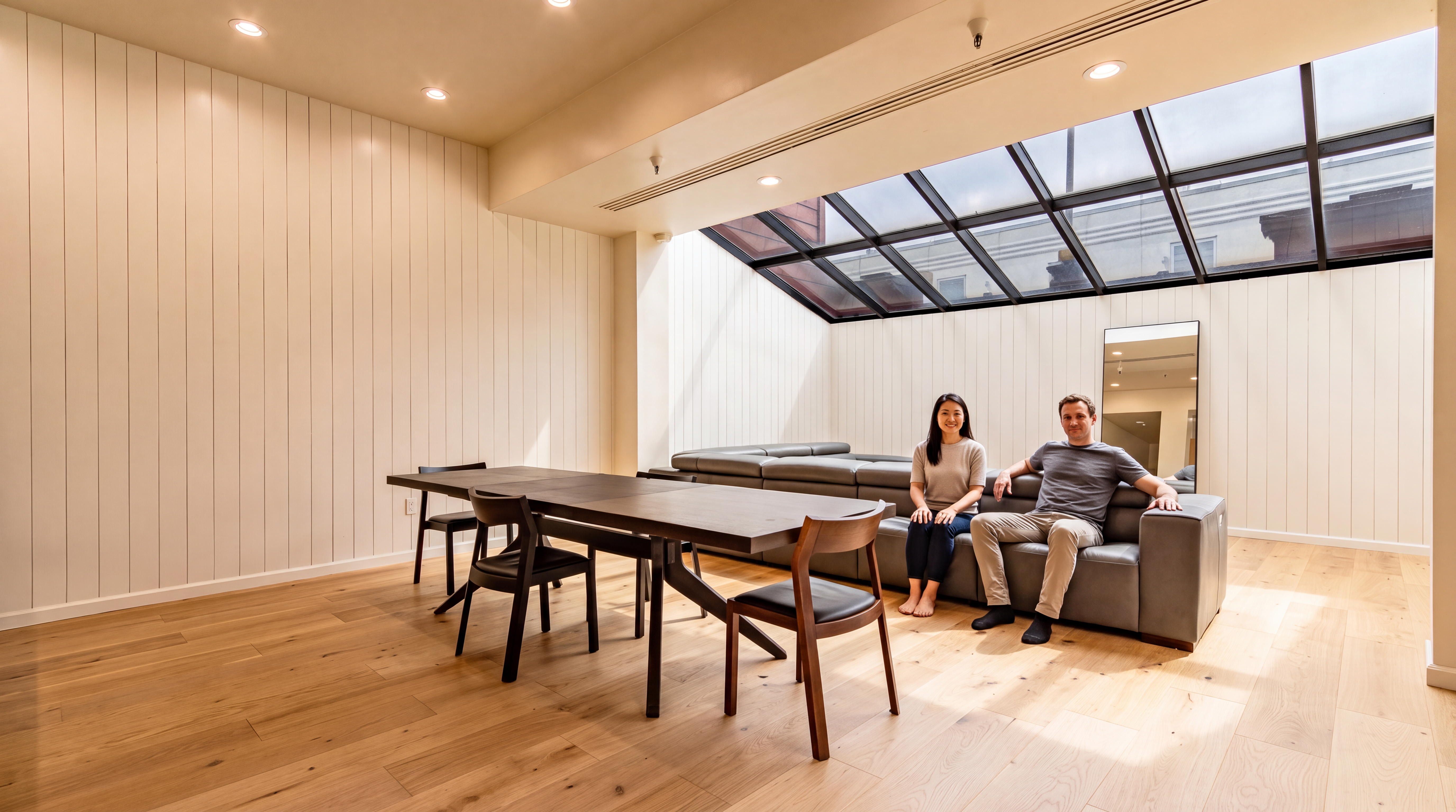 This modern, light-filled living area features a clean minimalist design with light wood flooring, vertical paneling, and an expansive sloped skylight that floods the room with natural light. The space is anchored by a sleek dark grey sectional sofa and a dark wood dining table, creating an open-concept multifunctional living and dining area. The aesthetic is contemporary and inviting, emphasizing an airy, architecturally interesting environment.