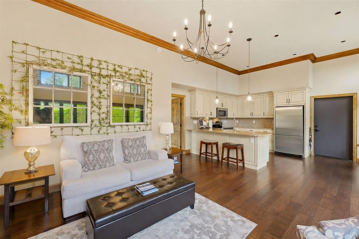 This open-concept living area features a cozy seating arrangement with a light-colored sofa and a dark leather ottoman, set against a wall decorated with two large window-style mirrors framed by climbing greenery. The space flows seamlessly into a kitchen equipped with light-toned cabinetry, a stone-topped island with bar seating, and stainless steel appliances. A large, multi-armed chandelier hangs from the high ceiling, casting a warm glow over the dark wood flooring that spans the entire room.