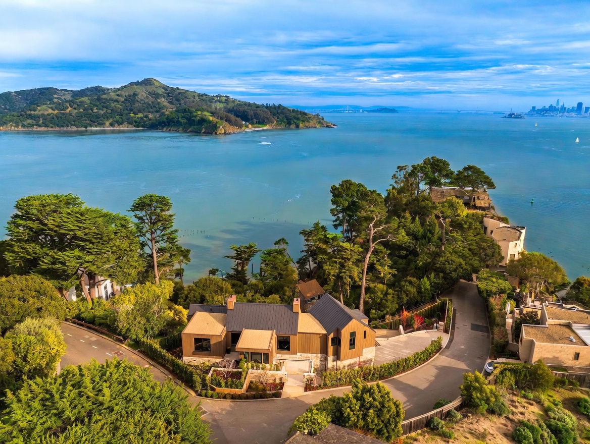 This high-angle aerial shot captures a modern, multi-level residence nestled on a lush, tree-lined coastal cliff overlooking a vast bay. The home features a dark metal roof, warm-toned wood siding, and stone accents, complemented by a landscaped courtyard and winding driveway. In the distance, the panoramic view includes a prominent island, a bridge, and the distant skyline of a major city under a bright, partly cloudy sky.