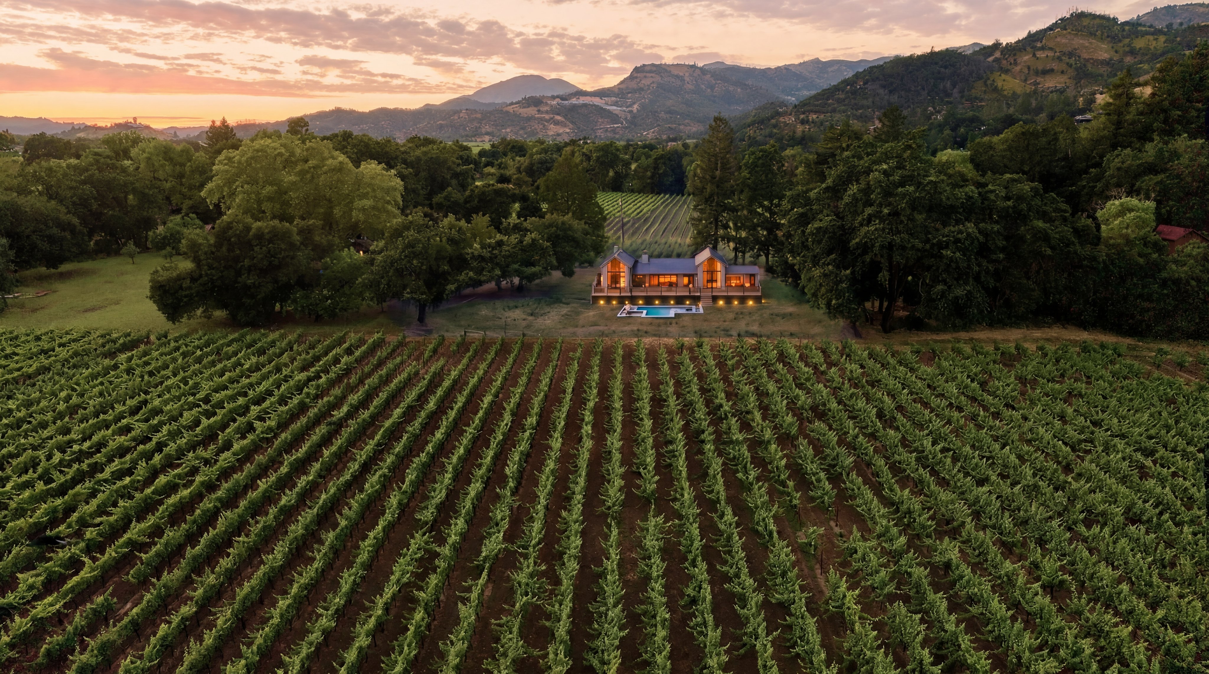 This stunning sunset aerial shot showcases a contemporary luxury home set amidst vast, neatly aligned vineyard rows. The residence features warm interior lighting, a spacious deck, and an inviting swimming pool, creating a serene and sophisticated atmosphere. The property is seamlessly integrated into the natural landscape, surrounded by lush trees and dramatic mountainous scenery.