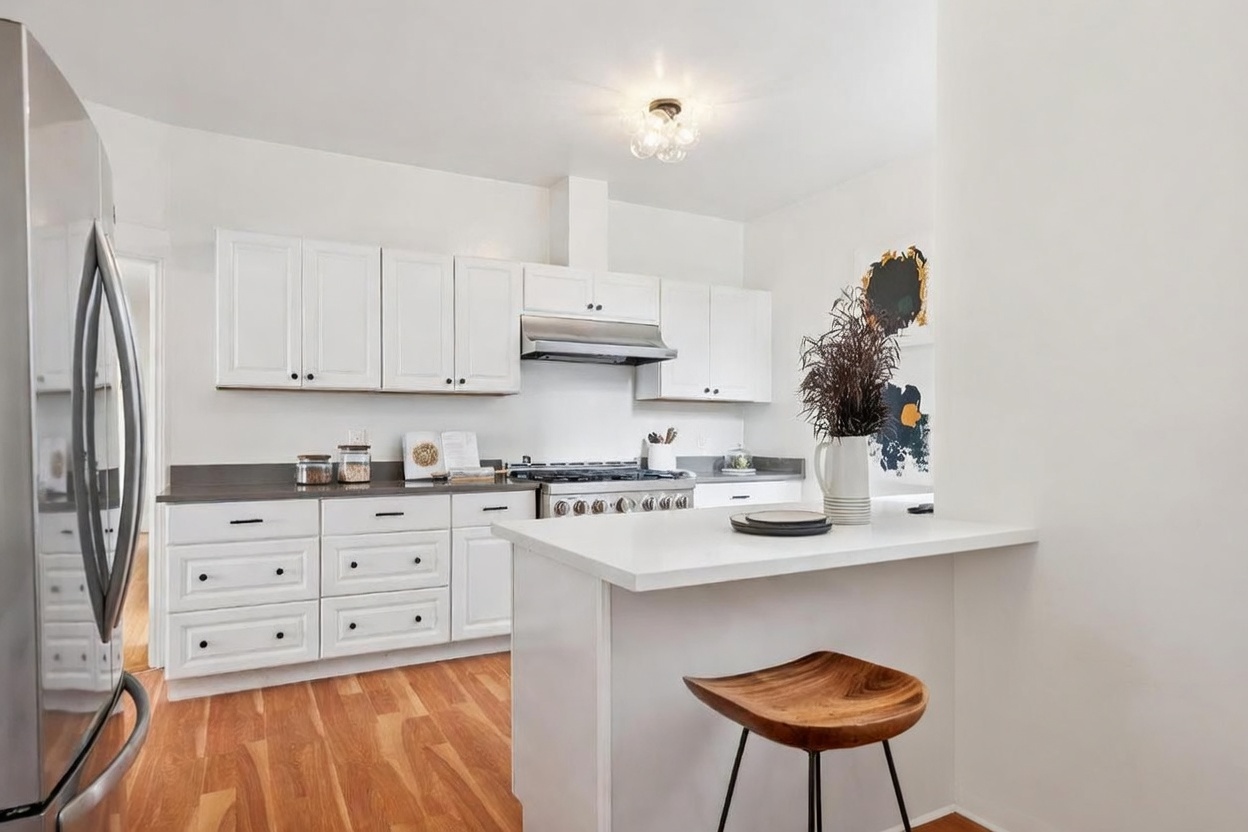 This bright, modern kitchen features white cabinetry, a stainless steel range with a matching hood, and a central island with a white countertop and two wooden bar stools. The space is accented by light wood flooring and a window that allows natural light to fill the room. The clean, minimalist aesthetic is complemented by a decorative light fixture and a simple, functional layout.