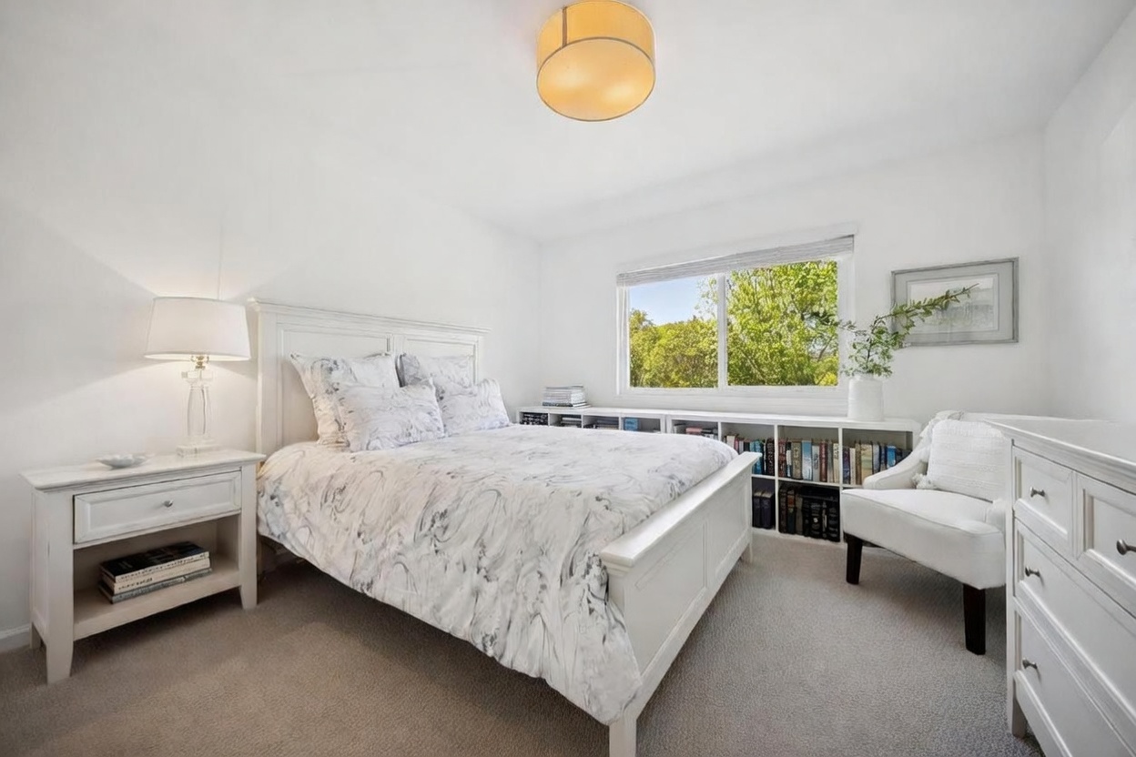 This bright and airy bedroom features a white bed frame with a patterned duvet, complemented by a matching white nightstand and a cozy armchair in the corner. A long, low bookshelf sits beneath a large window that provides ample natural light and a view of the surrounding greenery. The room is finished with neutral carpeting and a simple, warm-toned drum pendant light, creating a clean and inviting atmosphere.