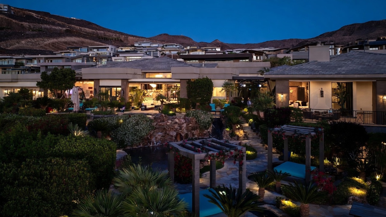 This dusk-lit exterior view captures a luxurious multi-level residence with a sprawling backyard oasis. The scene features a tiered landscape with a prominent rock waterfall, a stone-paved patio area with a pergola, and lush tropical greenery. The home's architecture is highlighted by warm ambient lighting, showcasing large glass openings that connect the indoor living spaces to the meticulously designed outdoor environment.