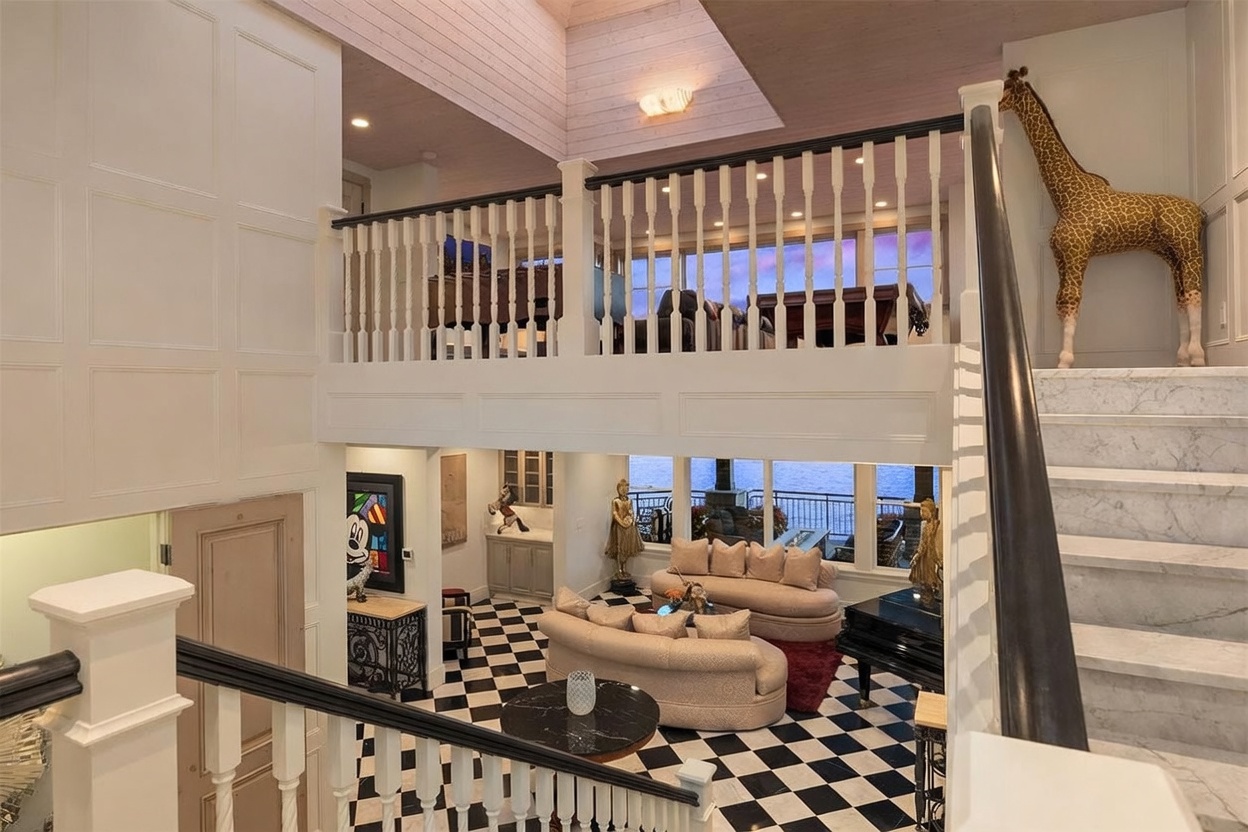 This grand, multi-level foyer features a dramatic staircase with a dark handrail and white balusters, overlooking a sunken living area with a striking black-and-white checkered floor. The space is characterized by high ceilings, a large giraffe sculpture, and expansive windows that provide a clear view of the water beyond. The open-concept design creates a luxurious, airy atmosphere that connects the upper landing to the main living space below.