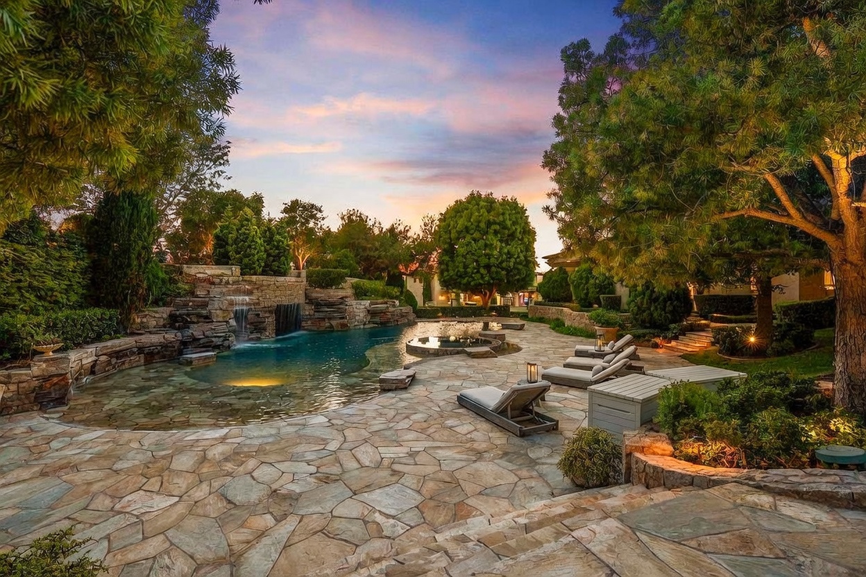 This image showcases a luxurious backyard pool and spa area at dusk. The pool features a natural stone waterfall and is surrounded by lush landscaping, including mature trees and manicured shrubs. Lounge chairs are arranged on the stone patio, creating an inviting and relaxing outdoor living space.