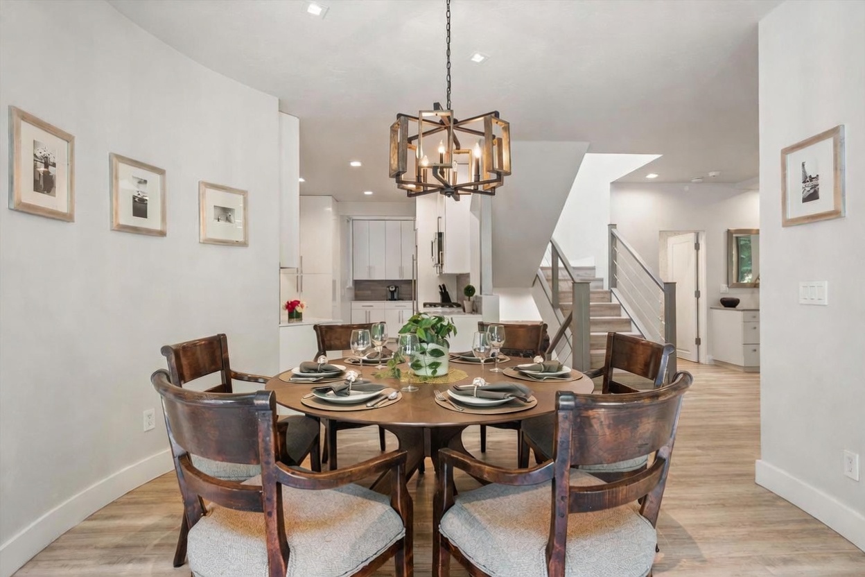 This dining room features a round wooden table surrounded by six upholstered chairs, positioned directly beneath a prominent geometric chandelier. The space is open-concept, flowing seamlessly into a bright kitchen area and a staircase with a modern railing. The neutral color palette and light wood-toned flooring create a clean, inviting atmosphere suitable for both casual and formal dining.
