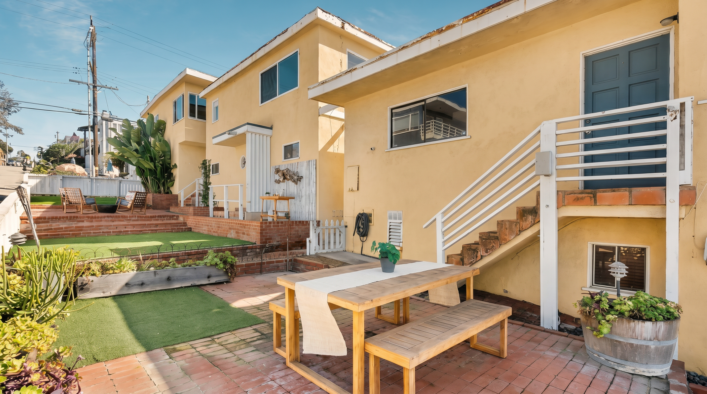 This outdoor space features a brick-paved patio area furnished with a light wood dining table and benches, perfect for al fresco meals. The scene includes a tiered yard with artificial turf, a small fire pit area, and a white staircase leading to an upper-level entrance. The warm-toned stucco exterior of the building and the surrounding greenery create a welcoming and relaxed atmosphere.
