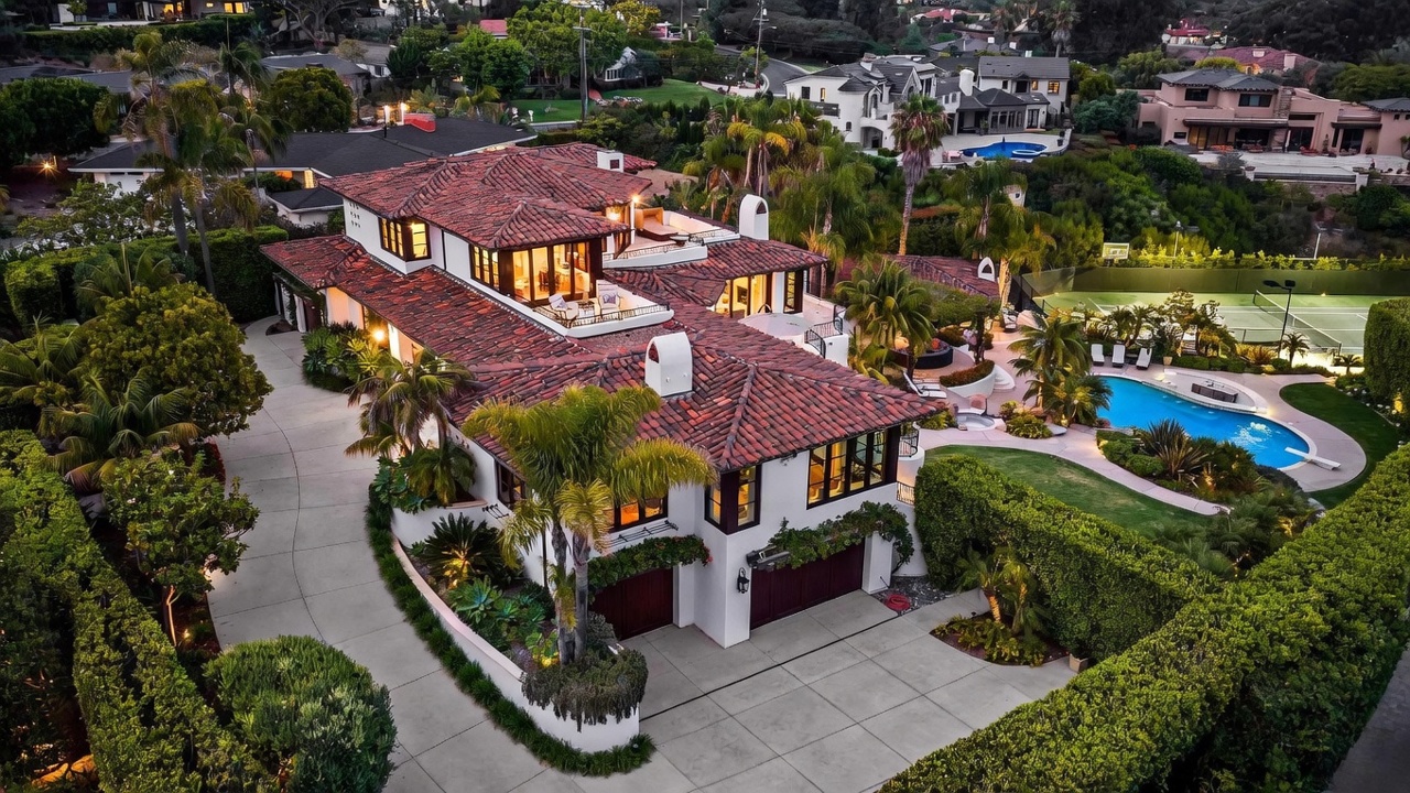 This high-angle aerial view captures a sprawling Mediterranean-style estate featuring a distinctive red-tiled roof and white stucco exterior. The property includes a private tennis court, a curved swimming pool, and lush, manicured landscaping surrounded by tall privacy hedges. The perspective highlights the expansive driveway, multiple outdoor living levels, and the seamless integration of luxury amenities within a secluded residential setting.