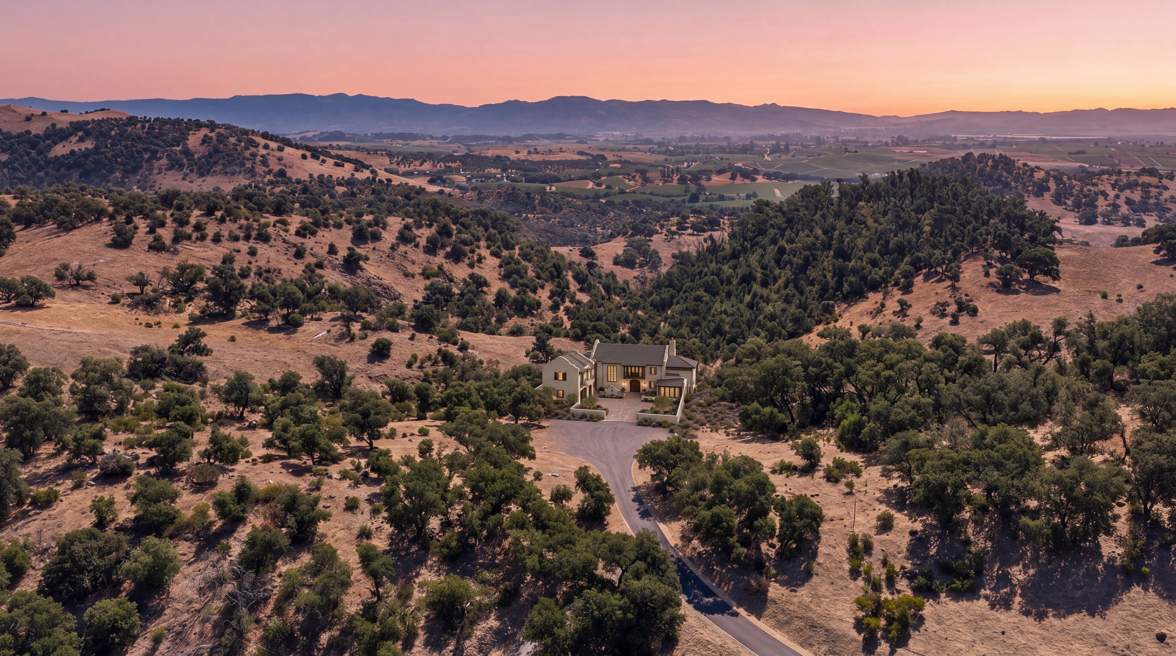This stunning aerial view captures a sophisticated Mediterranean-style estate nestled within a picturesque, rolling landscape of golden hills and mature oak trees. The property features a multi-gabled roofline, light-colored stucco exterior, and a gated courtyard entrance, set against the backdrop of a serene sunset. It offers a sense of privacy and tranquility, perfectly integrating the elegant architecture with its expansive rural environment.