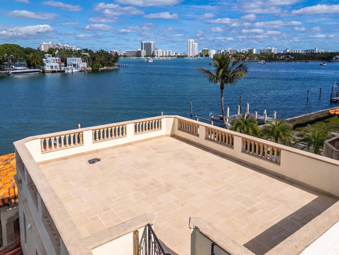 This elevated rooftop terrace features light-toned stone pavers and a classic balustrade railing, offering an expansive, unobstructed view of the bay and distant city skyline. The open-air space provides a serene vantage point overlooking the water, complete with a private dock and lush palm trees visible below. The perspective is captured from a slightly elevated angle, emphasizing the property's prime waterfront location and outdoor living potential.