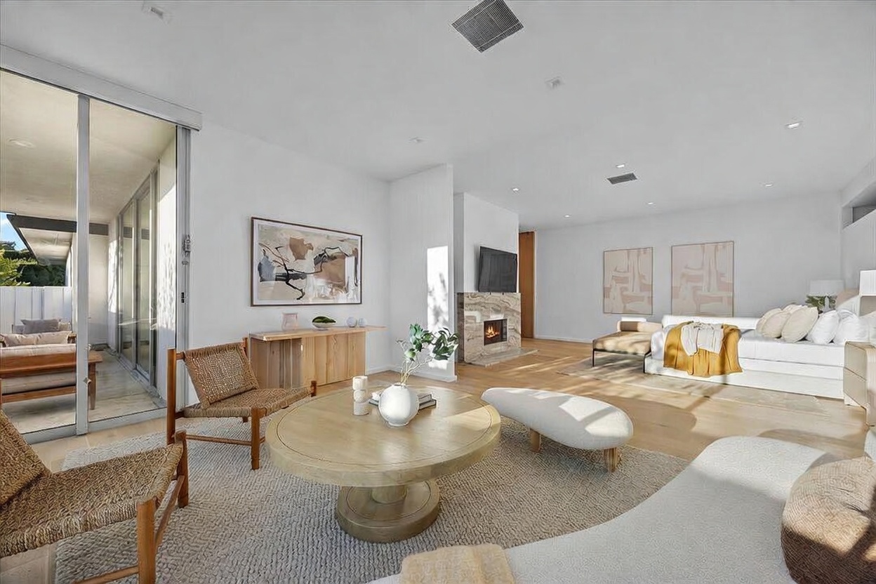 This expansive primary bedroom features a seamless transition between a cozy seating area and the sleeping quarters. The room is anchored by a stone-clad chimney breast with a fireplace and is illuminated by natural light streaming through large glass sliding doors that open to an outdoor patio. The neutral color palette, light wood flooring, and contemporary furnishings create a serene and sophisticated atmosphere.