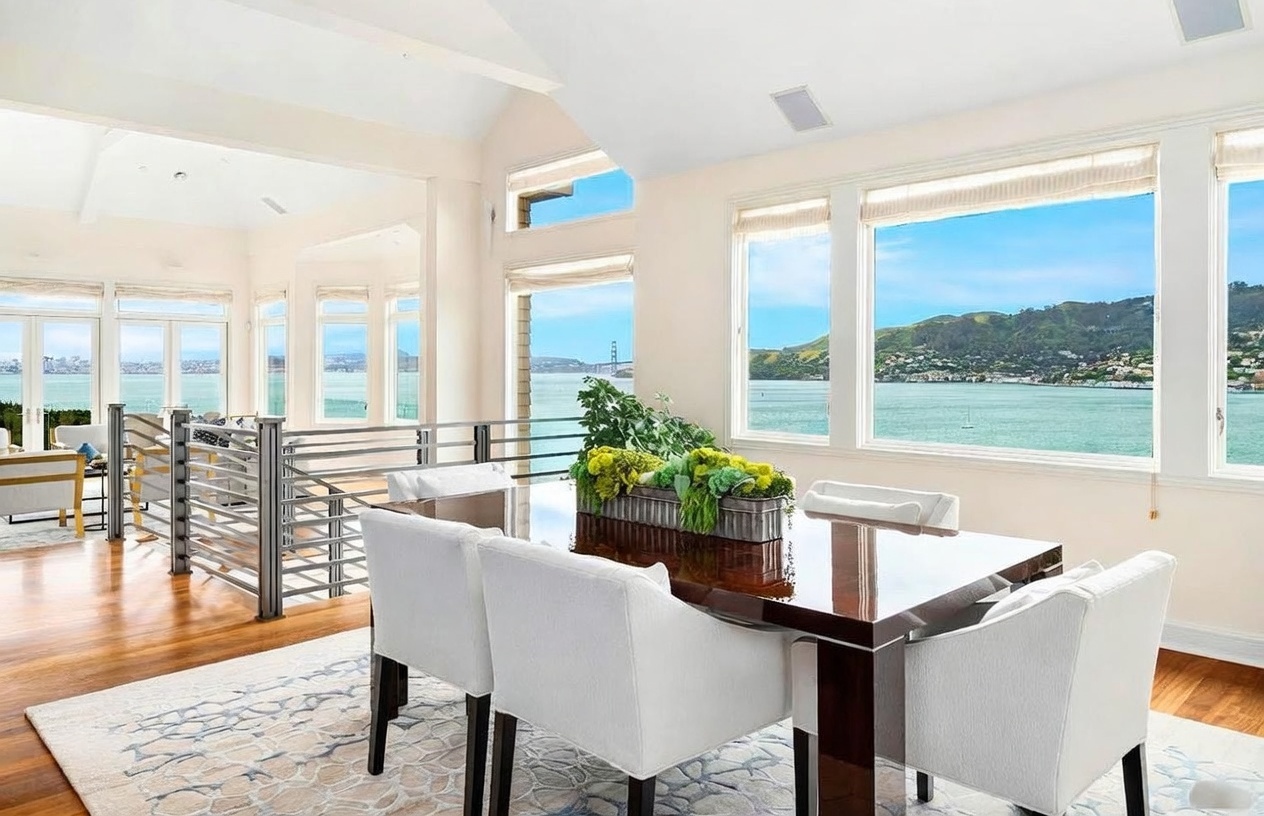 This bright and airy dining room features a dark wood table surrounded by white upholstered chairs, set atop a light-colored patterned area rug. Large windows provide expansive, panoramic views of the bay and distant hills, creating a serene and sophisticated atmosphere. The space is open-concept, with a metal railing overlooking a staircase and hardwood flooring that extends throughout the room.