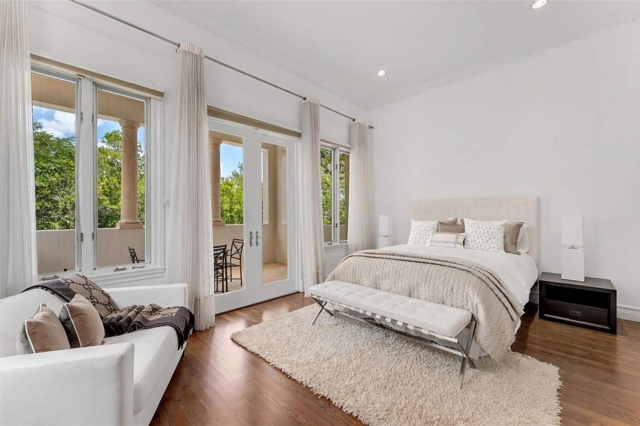 This bright and airy primary bedroom features a neutral color palette, hardwood flooring, and a plush area rug. The room is furnished with a tufted headboard bed, a white bench at the foot of the bed, and a white sofa in the foreground, all illuminated by natural light streaming through large windows and glass doors that lead to a balcony. The space offers a serene and sophisticated atmosphere with clean lines and elegant window treatments.