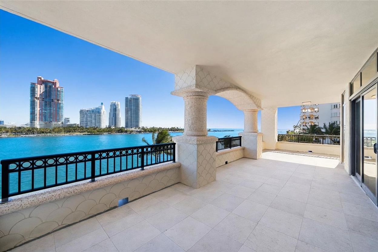This expansive, covered balcony features light-colored stone flooring and decorative, textured columns that frame a panoramic view of the bay and city skyline. The space is enclosed by a black metal railing, offering a seamless transition between the indoor living area and the outdoor environment. The bright, sunny atmosphere highlights the prime waterfront location and the generous proportions of the outdoor living space.