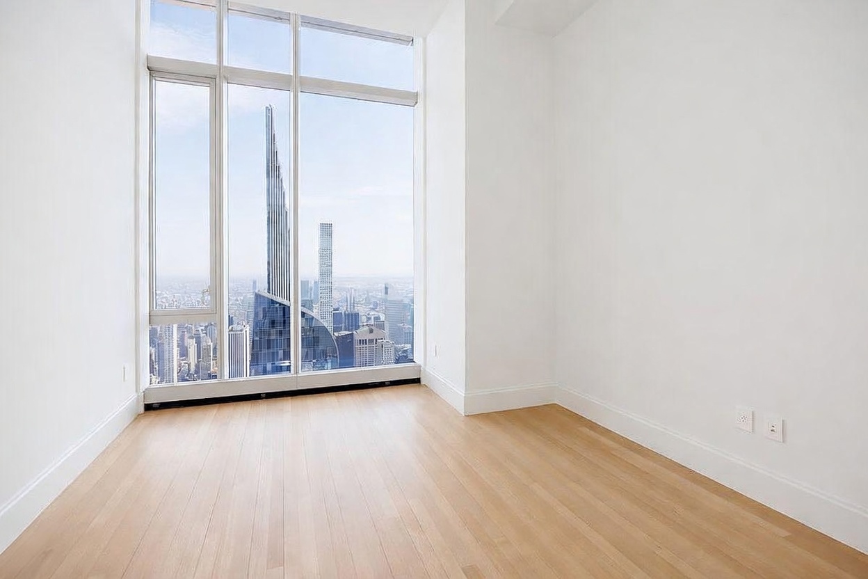 This minimalist room features light wood flooring and crisp white walls, creating a bright and airy atmosphere. A large, floor-to-ceiling window assembly dominates the space, offering a dramatic, unobstructed view of the surrounding city skyline. The perspective is centered on the window, emphasizing the height and urban connection of the property.