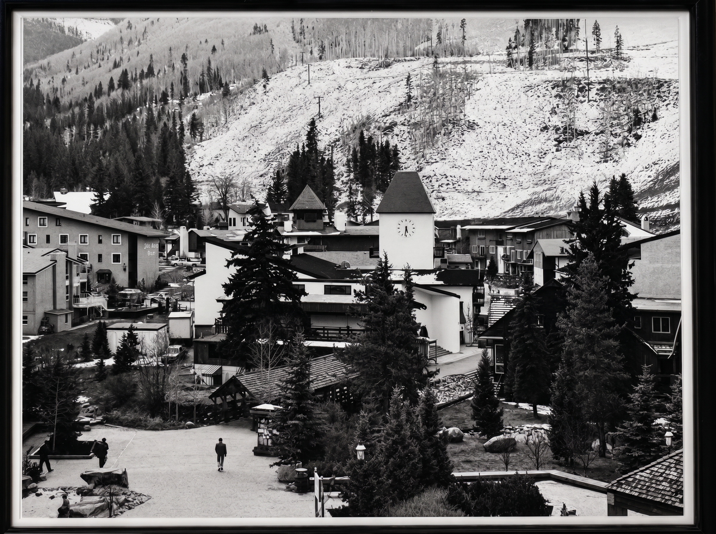 This black and white photograph captures a village-style resort setting nestled at the base of a snow-dusted, tree-lined mountain. The scene features a central clock tower, pedestrian walkways, and mixed-use buildings surrounded by evergreen landscaping, evoking a charming, alpine-inspired atmosphere. It highlights a walkable, master-planned community layout perfect for a scenic ski-resort or vacation village environment.