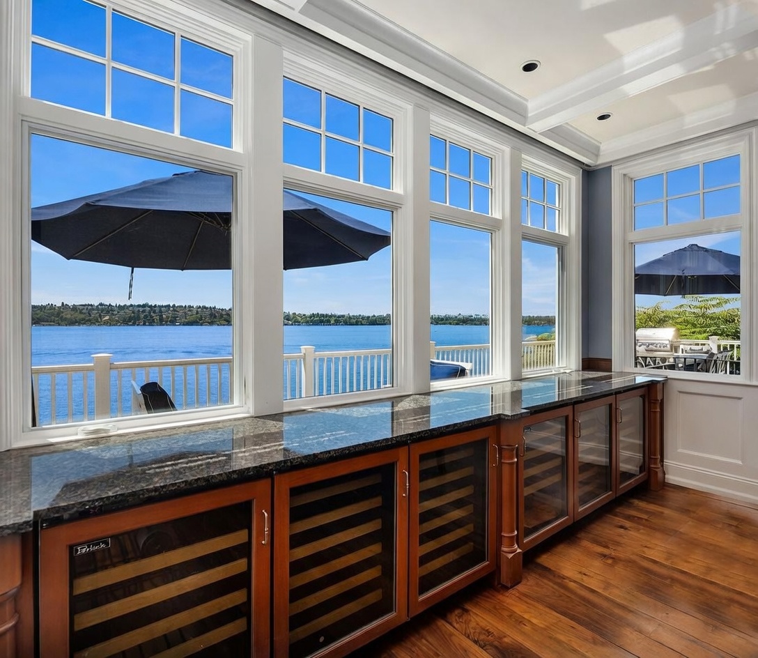 This bright and airy sunroom features a long, custom-built wet bar with dark granite countertops and multiple under-counter wine refrigerators. Large, multi-pane windows provide an expansive, panoramic view of the lake and outdoor deck, creating a seamless connection between indoor luxury and the natural landscape. The rich hardwood flooring and elegant cabinetry contribute to a sophisticated, relaxing atmosphere perfect for entertaining.
