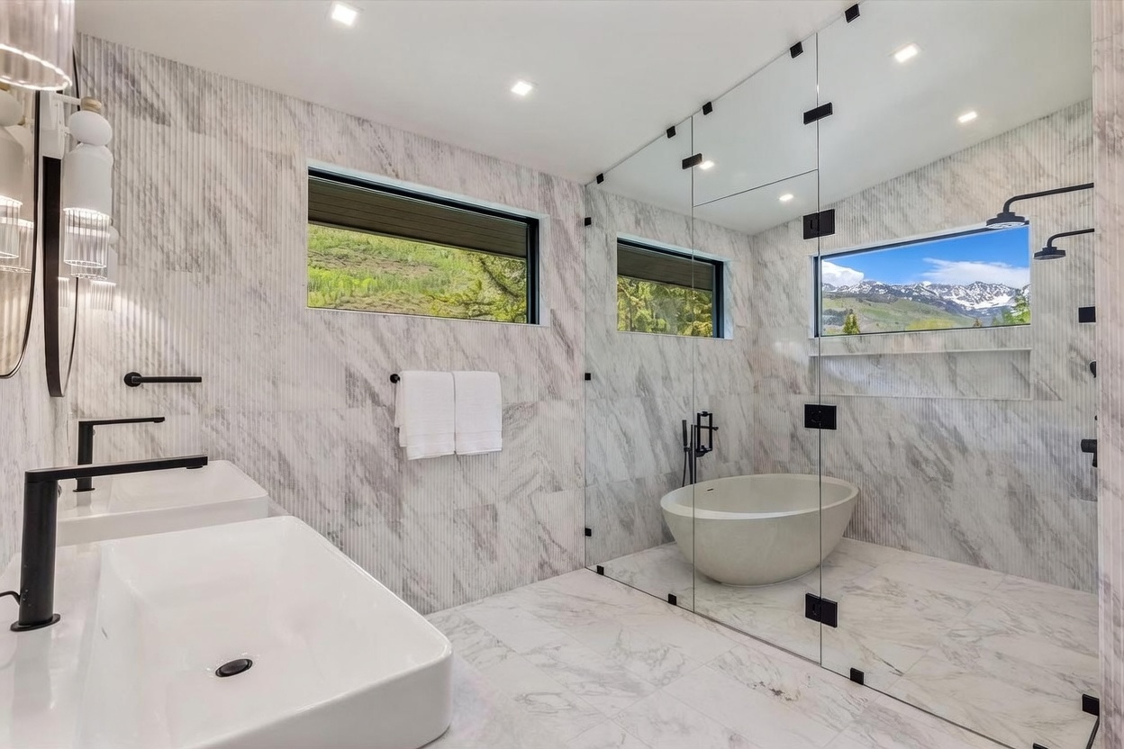 This luxurious primary bathroom features a sleek, modern design with extensive stone wall and floor tiling. A glass-enclosed wet room houses a freestanding soaking tub and a shower area, while a minimalist vanity with a black faucet sits to the left. Large windows provide natural light and scenic mountain views, creating a serene and high-end atmosphere.