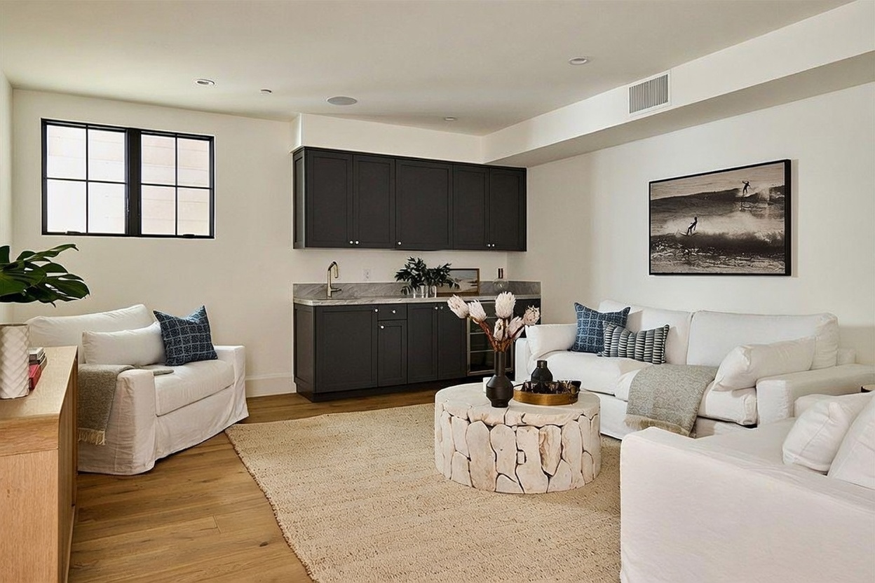 This bright and inviting living room features a neutral color palette with comfortable white seating arranged around a textured, circular stone coffee table. A built-in wet bar with dark cabinetry and a stone countertop provides a sophisticated focal point against the back wall. The space is accented by a large black-and-white surf-themed wall art piece and light wood flooring, creating a relaxed, coastal-inspired atmosphere.