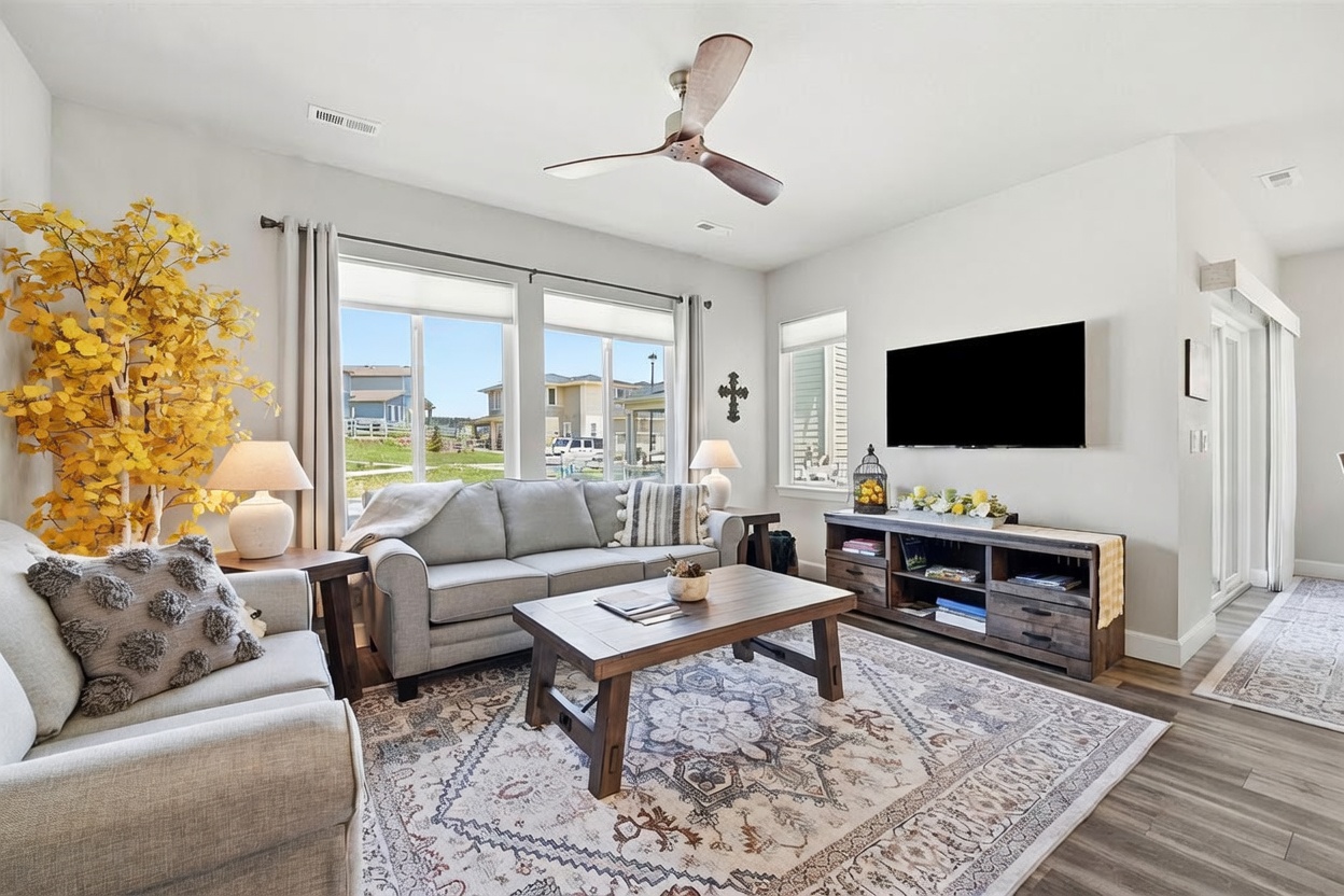 This bright and inviting living room features a neutral color palette, light-toned walls, and wood-look flooring. The space is furnished with a comfortable gray sofa, a rustic wooden coffee table, and a media console topped with a television. Large windows allow for ample natural light, while a decorative tree and a patterned area rug add texture and warmth to the room.