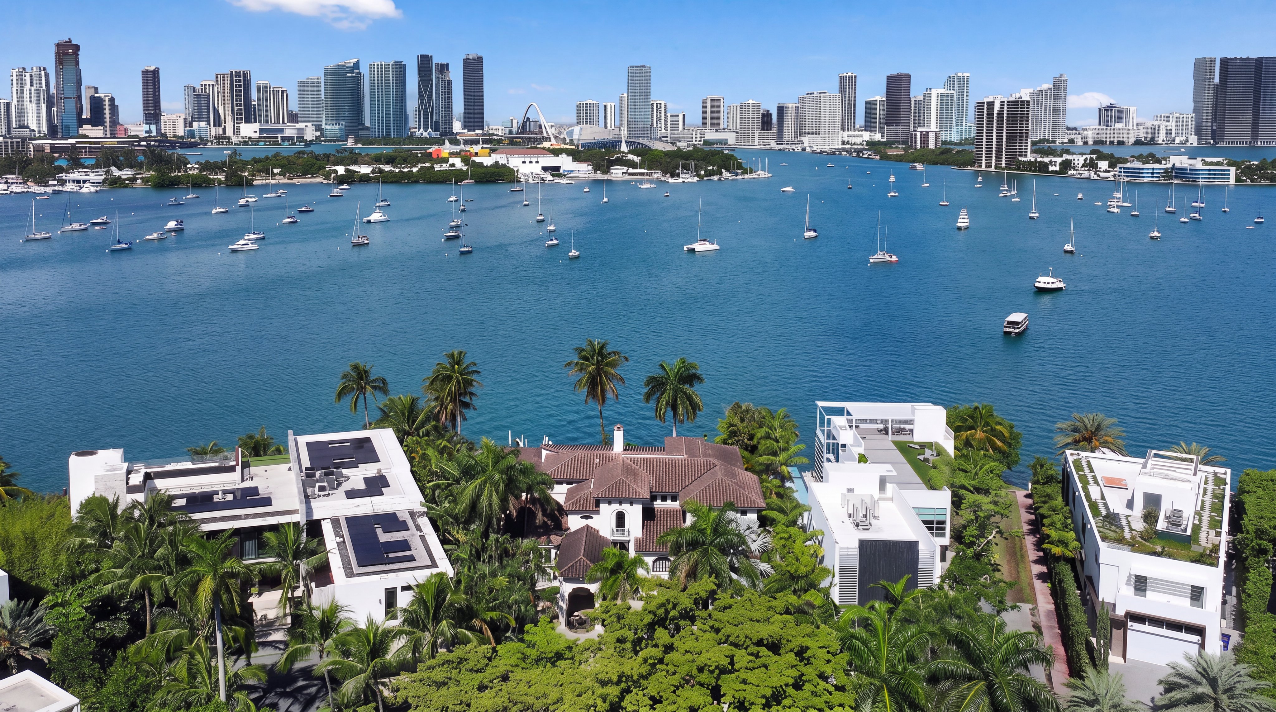 This stunning aerial view captures a cluster of luxury waterfront properties featuring modern architectural designs, flat roofs with solar panels, and lush tropical landscaping. The homes overlook a vibrant bay filled with numerous sailboats and yachts, set against the impressive metropolitan skyline of Miami. This prime location offers a blend of tranquil waterfront living and convenient access to the bustling city center.