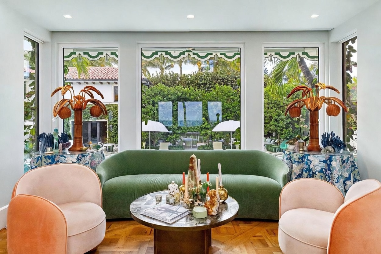 This elegant living room features a curved green velvet sofa centered in front of three large windows that provide a view of a lush, green garden. Two soft-toned, rounded armchairs flank a circular stone-topped coffee table, while decorative palm-shaped lamps sit on patterned tables on either side. The room is anchored by a warm, herringbone-patterned wood floor, creating a sophisticated and inviting atmosphere.