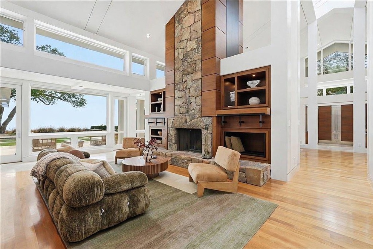 This expansive living room features a dramatic stone-clad chimney breast that serves as the central focal point, flanked by custom wood cabinetry and shelving. The space is bathed in natural light from large windows that offer a view of the outdoors, complemented by light wood flooring and a textured area rug. The room is furnished with a plush, rounded sofa and two accent chairs, creating a sophisticated and airy atmosphere.