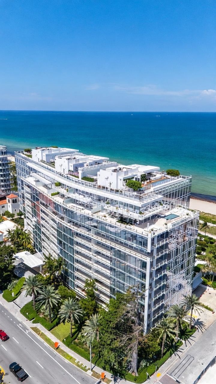This high-angle aerial shot captures a modern, multi-story luxury residential building situated directly along a sandy coastline. The structure features extensive glass facades, multiple terraced levels with private outdoor spaces, and a rooftop area that includes a pool. Lush tropical landscaping surrounds the base of the building, which is positioned adjacent to a coastal road with light vehicular traffic.