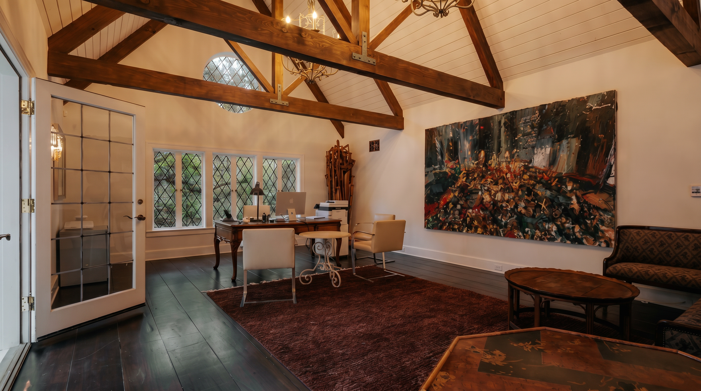 This sophisticated home office features dramatic vaulted ceilings with exposed wooden beams and a large, abstract art piece that serves as a focal point. The room is furnished with a classic wooden desk, modern chairs, and a plush dark rug, all set against dark hardwood floors. A large window with diamond-pane detailing allows natural light to fill the space, creating an elegant and professional atmosphere.