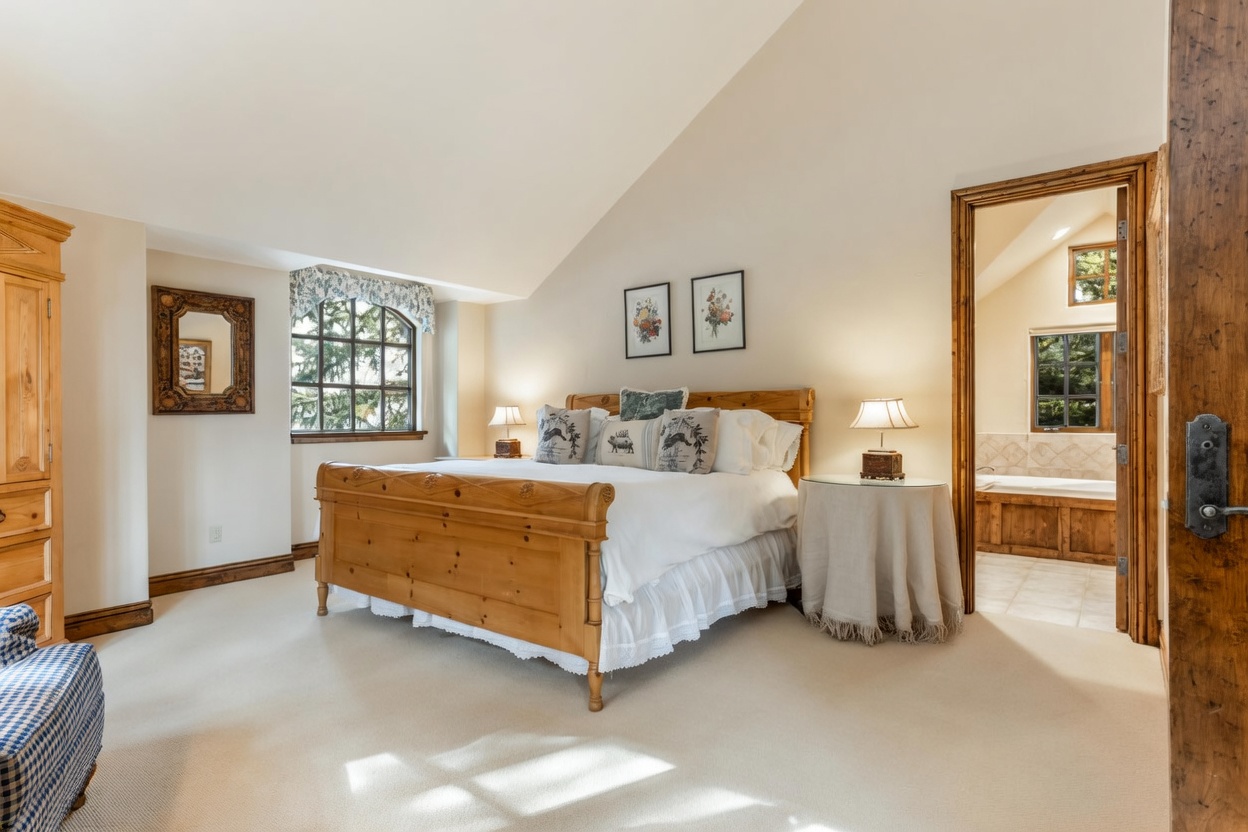 This spacious primary bedroom features a vaulted ceiling and a large wooden bed frame as the central focal point. The room is decorated in a neutral, traditional style with soft carpeting, a round side table with a lamp, and a framed mirror on the wall. A doorway leads into an adjoining bathroom, providing a cohesive and inviting layout.