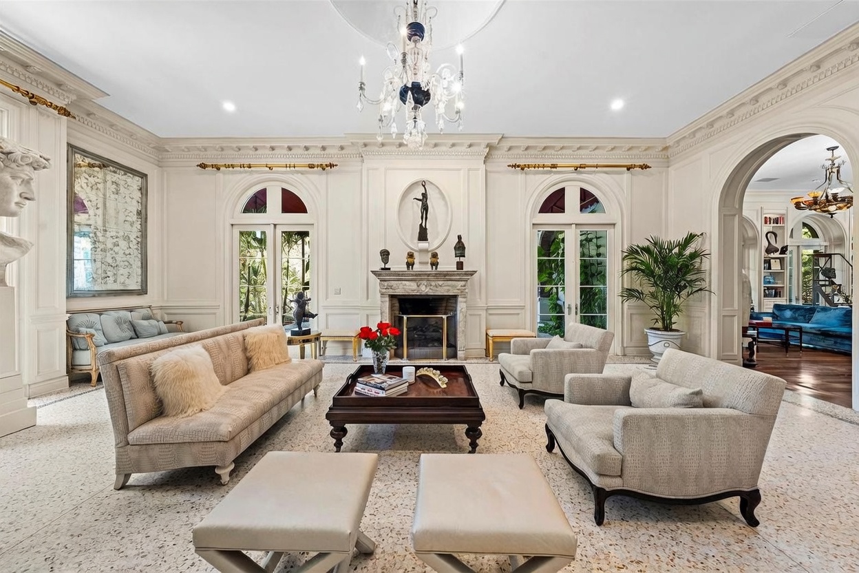 This elegant, formal living room features a symmetrical layout centered around a stone fireplace with a decorative mantel and a tall, ornate surround. The space is furnished with a plush sofa, two armchairs, and a pair of ottomans, all arranged on a light-colored, speckled stone floor. Large glass doors with arched tops flank the fireplace, allowing natural light to fill the room, while a grand crystal chandelier hangs from the center of the ceiling, adding a touch of luxury to the classic architectural details.