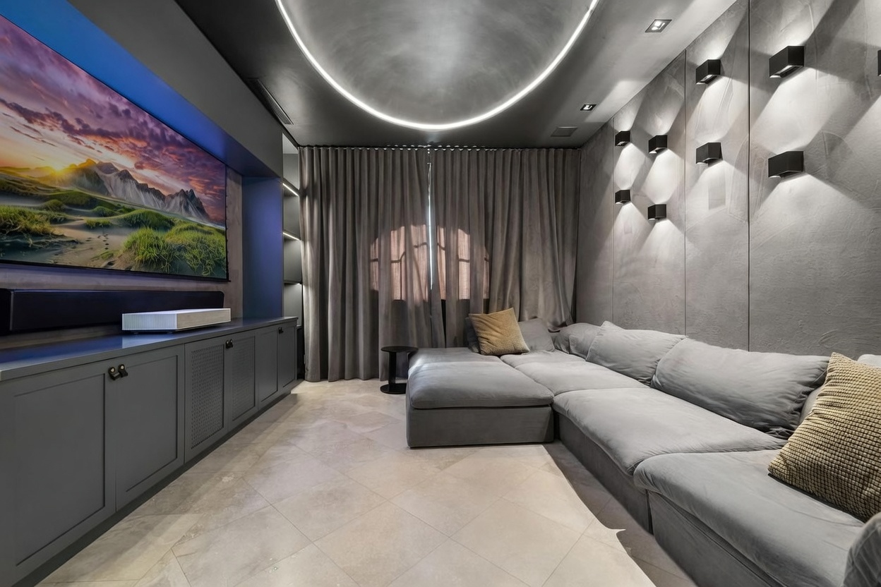 This modern media room features a large, plush grey sectional sofa arranged against a textured grey wall accented by geometric wall-mounted light fixtures. A large television is mounted above a sleek, dark-toned media console, while a curved LED light strip embedded in the ceiling provides a dramatic, contemporary glow. The room is finished with light-colored tile flooring and floor-length curtains that add a soft, cozy texture to the sophisticated space.