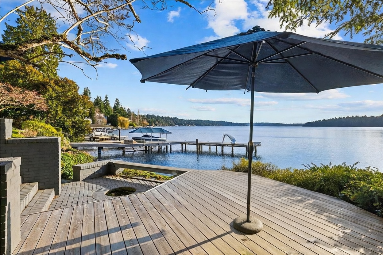 This scenic outdoor deck features a weathered wooden dining set shaded by a large dark umbrella, overlooking a serene lake. The perspective captures the inviting waterfront living space, complete with a private dock, boat lift, and a slide extending into the water. Lush trees and a clear blue sky frame the tranquil, picturesque setting, highlighting the property's prime lakefront location.