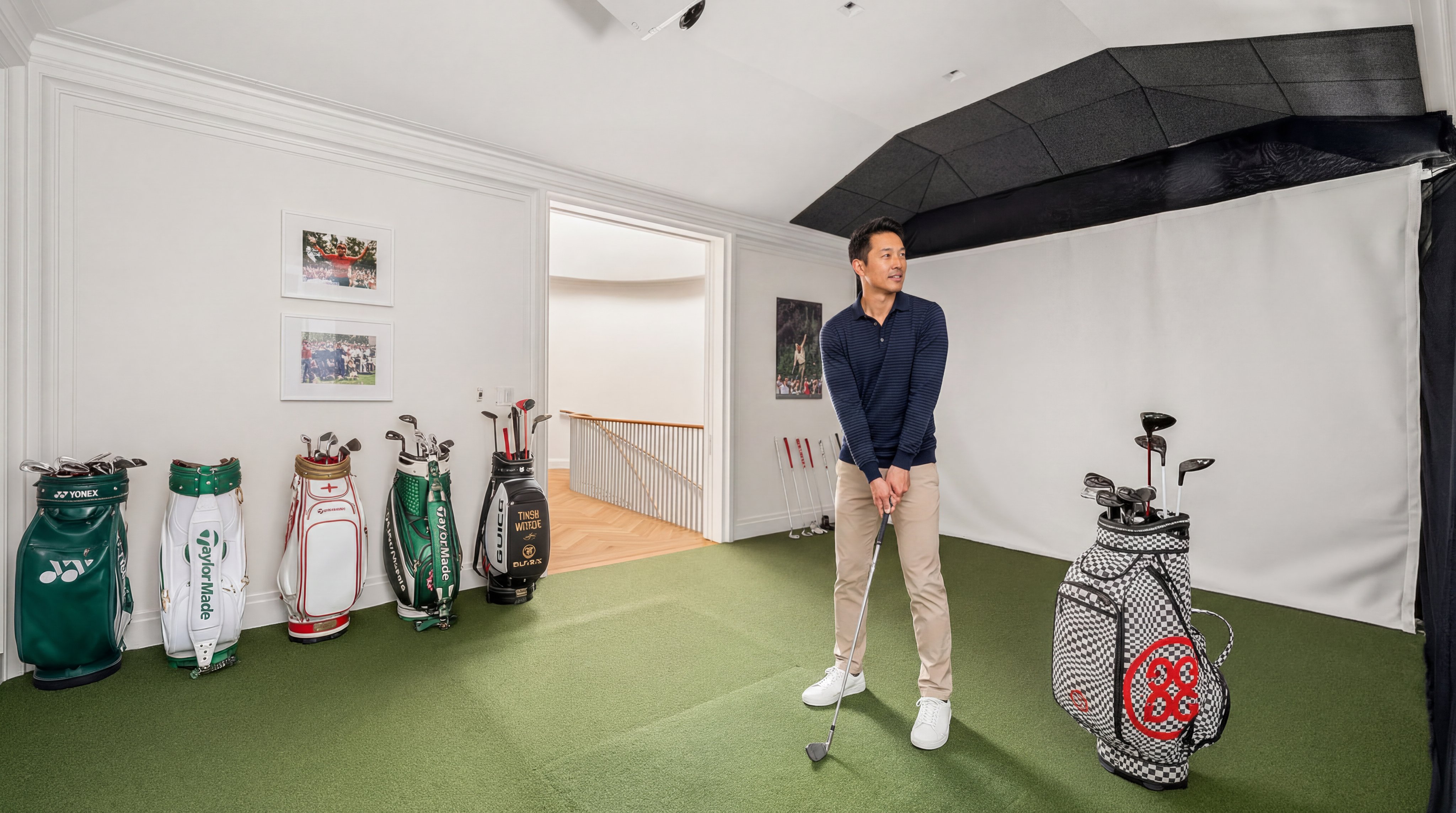 This dedicated indoor golf simulator room provides a professional practice space, featuring high-quality turf flooring, a large practice projection screen, and multiple decorative golf bags. The room is seamlessly finished with gallery-style wall art, crown molding, and an open doorway leading to a staircase, reflecting a luxurious and hobby-focused lifestyle amenity.