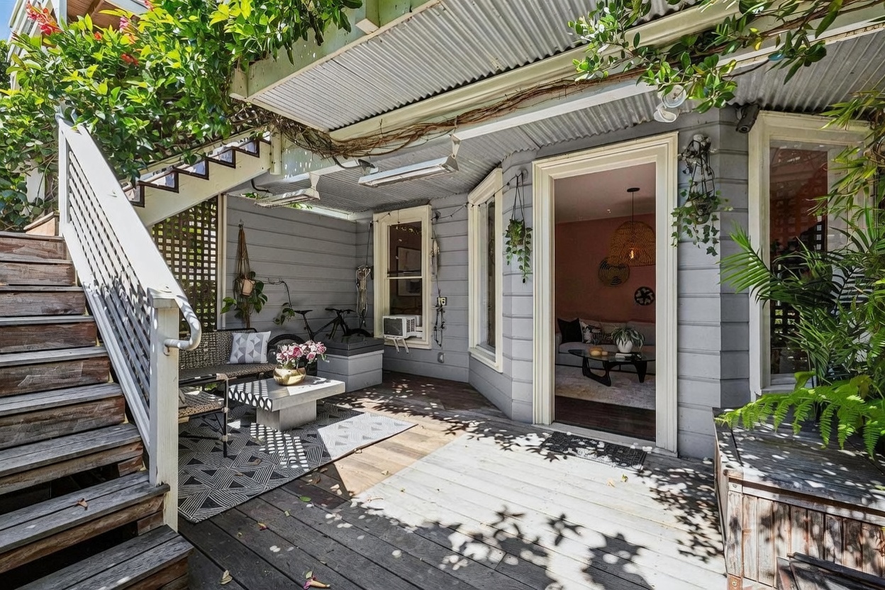 This inviting outdoor deck features a covered seating area with a patterned rug, a modern coffee table, and a bench, all nestled under a corrugated metal roof. A wooden staircase leads up to an upper level, while a doorway provides a seamless transition into a cozy, pink-toned living space visible inside. Lush greenery and climbing vines frame the deck, creating a private, tranquil atmosphere perfect for relaxation.