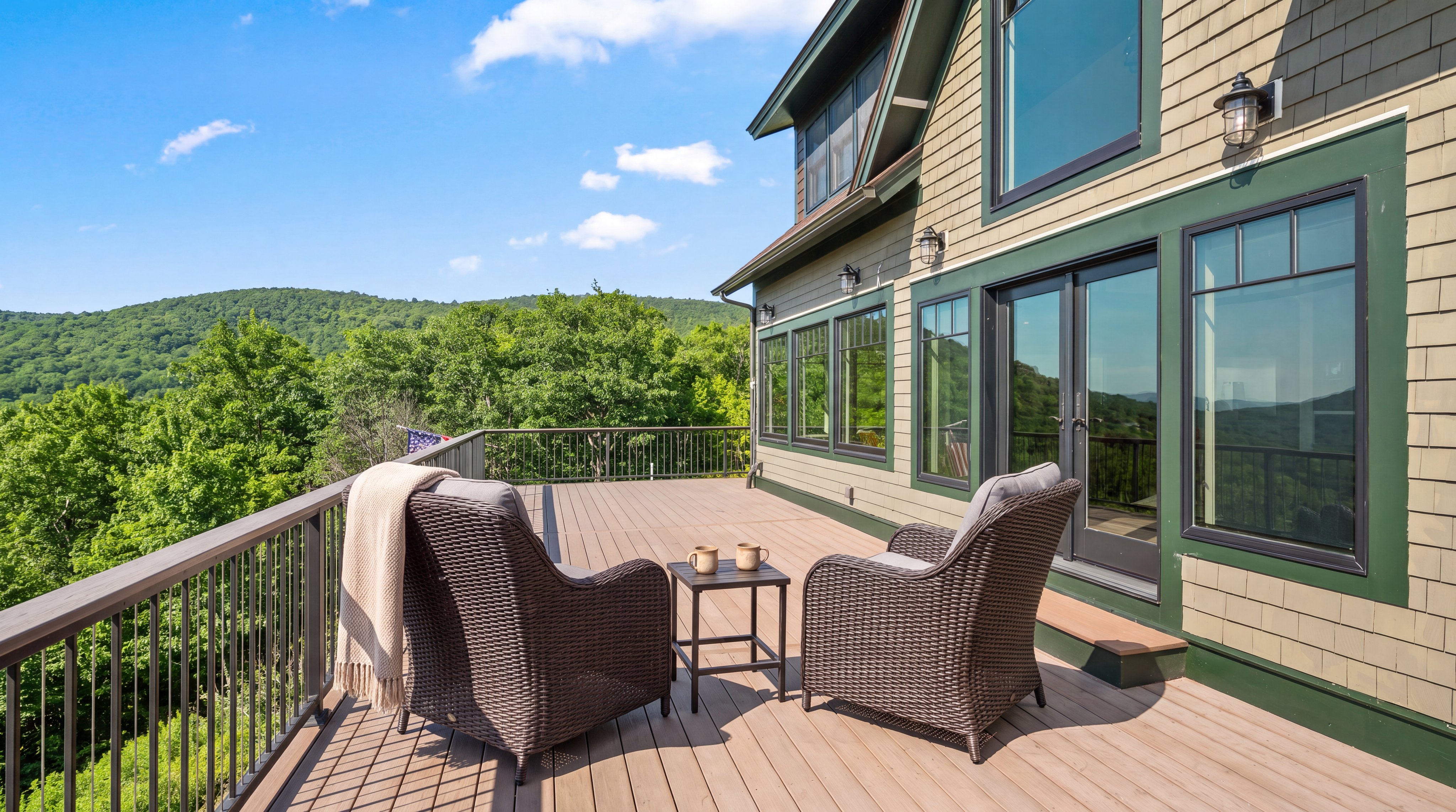 This spacious wooden deck features two comfortable wicker armchairs with a small centrale side table, creating a serene spot to enjoy the expansive mountain vistas. The home exhibits a charming cedar-shingle and green-trim architectural style, with large, bright windows overlooking the lush wooded landscape. It offers an inviting outdoor living space perfect for relaxation and capturing scenic panoramic views.
