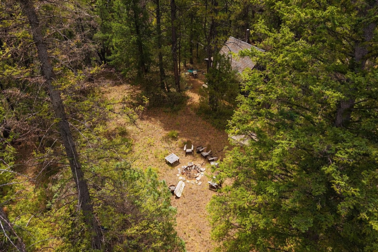 This high-angle aerial view captures a secluded cabin nestled within a dense, wooded forest. The property features a rustic outdoor fire pit area surrounded by wooden chairs, creating a peaceful and private retreat. The perspective highlights the cabin's integration into the natural landscape, emphasizing the serene and quiet atmosphere of the setting.