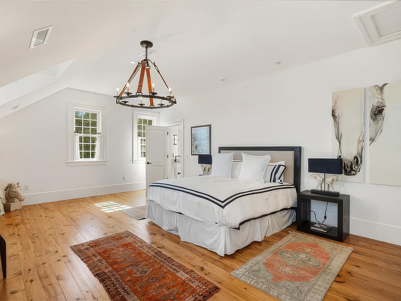 This spacious primary bedroom features light wood flooring and a vaulted ceiling, creating an airy and open atmosphere. A large bed with a dark frame serves as the focal point, flanked by two nightstands with black lamps and accented by large horse-themed artwork on the wall. The room is illuminated by a rustic-style chandelier and natural light streaming through the windows, while two patterned area rugs add texture to the floor.