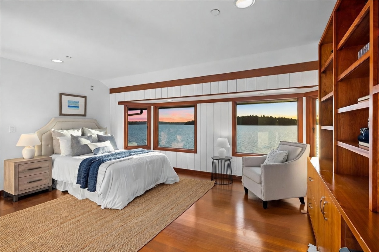 This serene primary bedroom features a neutral color palette, a plush bed with a blue throw, and a cozy armchair positioned to take advantage of the stunning waterfront views. The room is accented by warm wood trim, built-in shelving, and a natural fiber rug, creating a tranquil and inviting atmosphere. The perspective is wide-angle, capturing the seamless blend of comfortable indoor living and the expansive natural scenery outside.